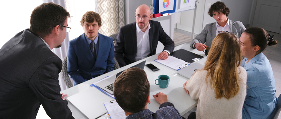 A diverse team gathered around a conference table in discussion, with notebooks, laptops, and charts visible in a meeting room.