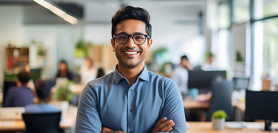 Smiling man with glasses in a blue shirt stands with arms crossed in a bright, open office.